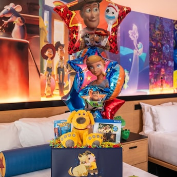 A Pixar Welcome Celebration decor package displayed on top of a bed features a gift bag overflowing with balloons, a Dug plush, a puzzle, a sweet treat and confetti